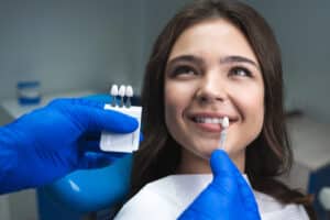 young woman sitting in dentist chair, smiling while dentist holds veneer up to her natural teeth