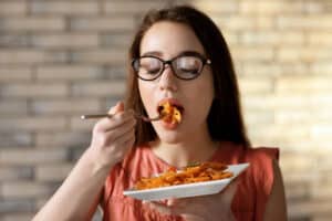 woman taking a bite of pasta, holding a white plate full of pasta