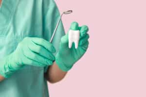 dentist in scrubs and gloves holding a large model tooth and dental mirror on pink background